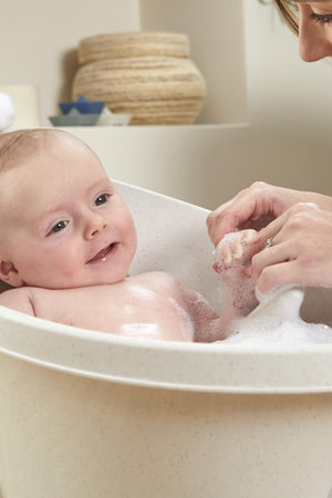 A smiling baby in a speckled white bathtub is being gently washed with bubbles by an adults hands.