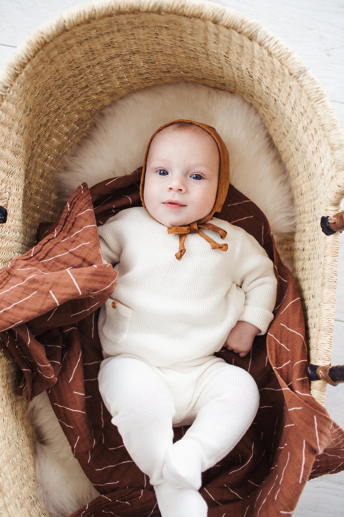 A baby in a cozy brown muslin swaddle and white outfit lies in a woven basket.