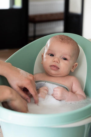 A baby in a mint green baby bath with supportive backrest, surrounded by bubbles and a guiding hand.