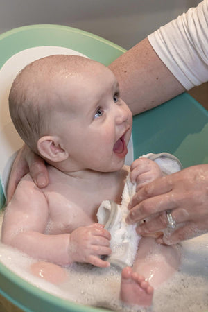 A smiling baby sits in a green and white bathtub, surrounded by bubbles, holding a washcloth.