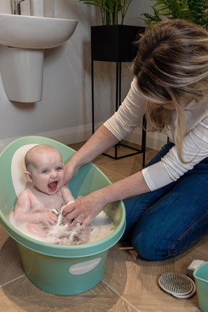 A smiling baby in a green Shnuggle baby bath with an adult gently washing, surrounded by bathroom decor.