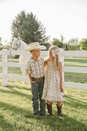 Two children stand together in a sunlit outdoor setting, wearing Western-themed outfits. The boy wears a patterned shirt and jeans, while the girl dons a pony print dress and boots.