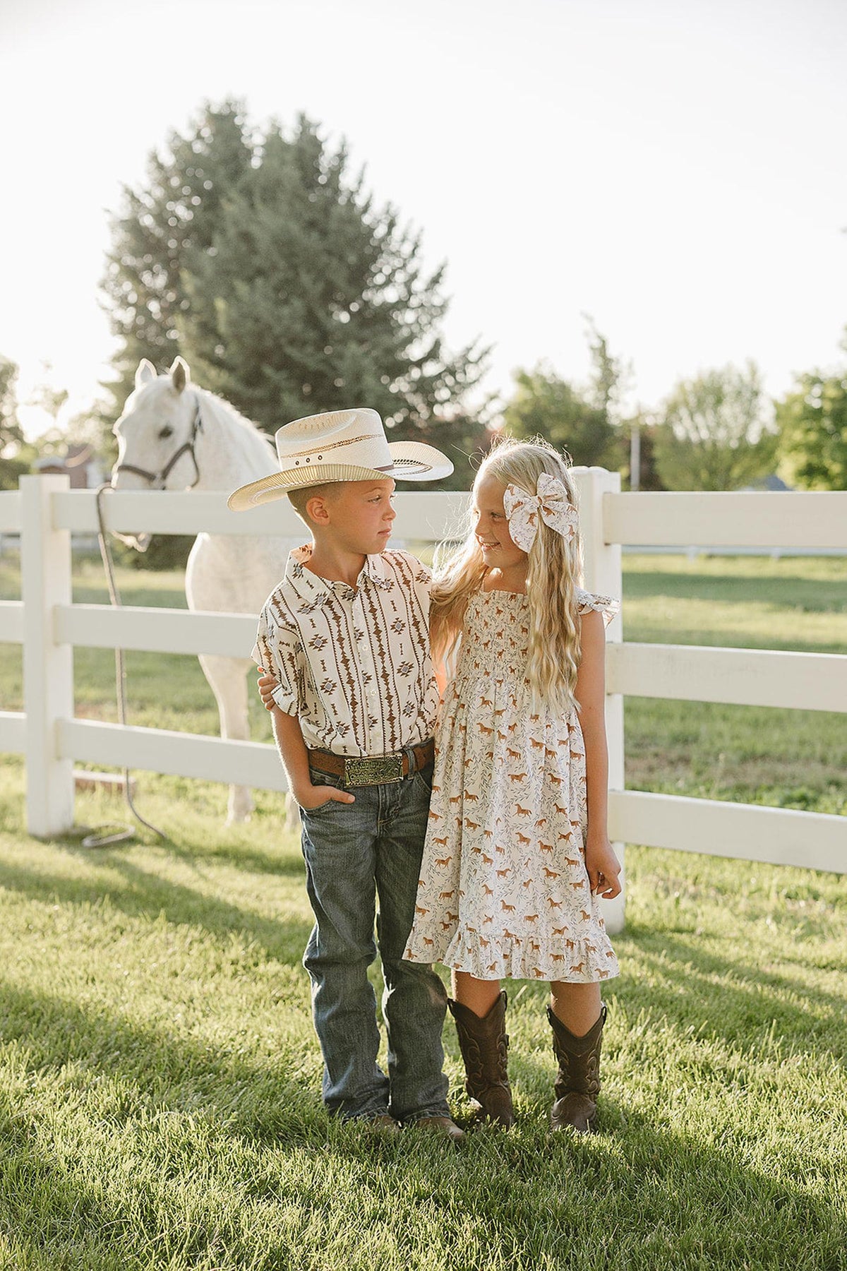 A young boy in a Western-style button-up shirt stands beside a girl in a floral dress, both wearing cowboy boots.