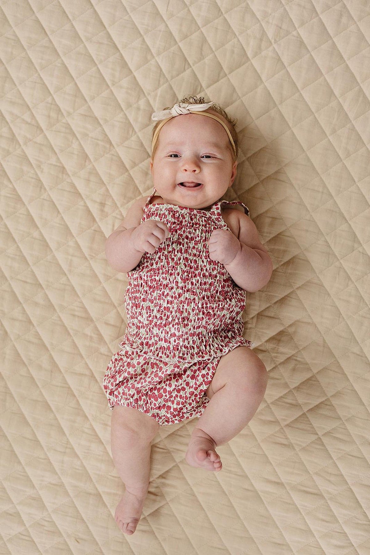 A smiling baby girl wears a floral ruched romper and a headband, lying on a quilted beige blanket.