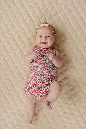 A smiling baby girl wears a floral ruched romper and a headband, lying on a quilted beige blanket.
