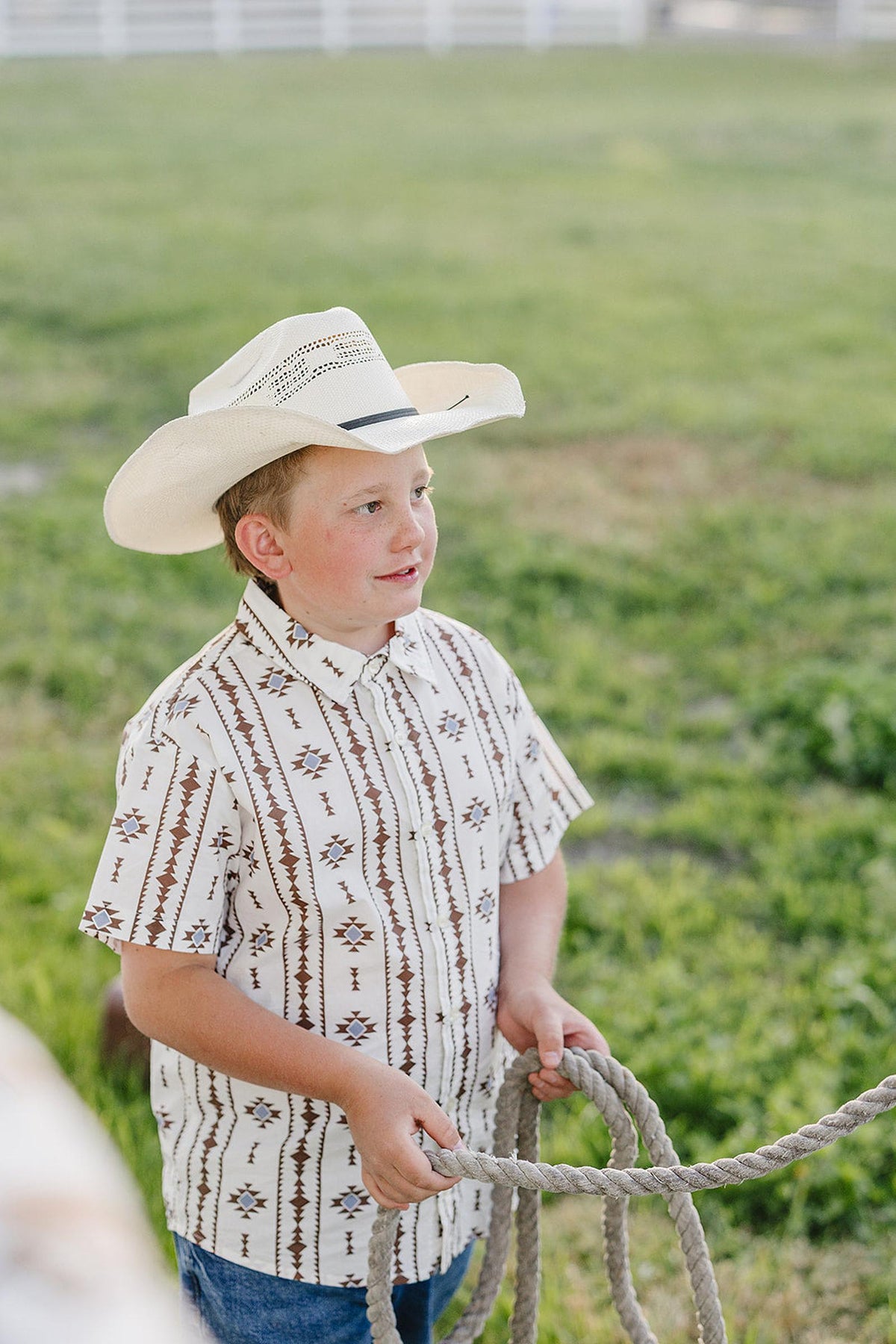 A young boy wearing a white cowboy hat and a patterned button-up shirt stands outdoors, holding a rope. The grassy background suggests a rustic, western setting ideal for adventures.