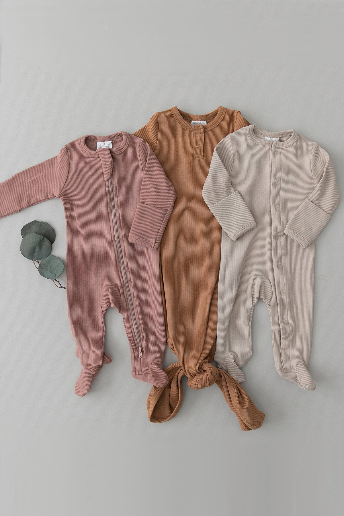 Three ribbed baby pajamas in dusty rose, brown, and beige are laid out on a light surface.