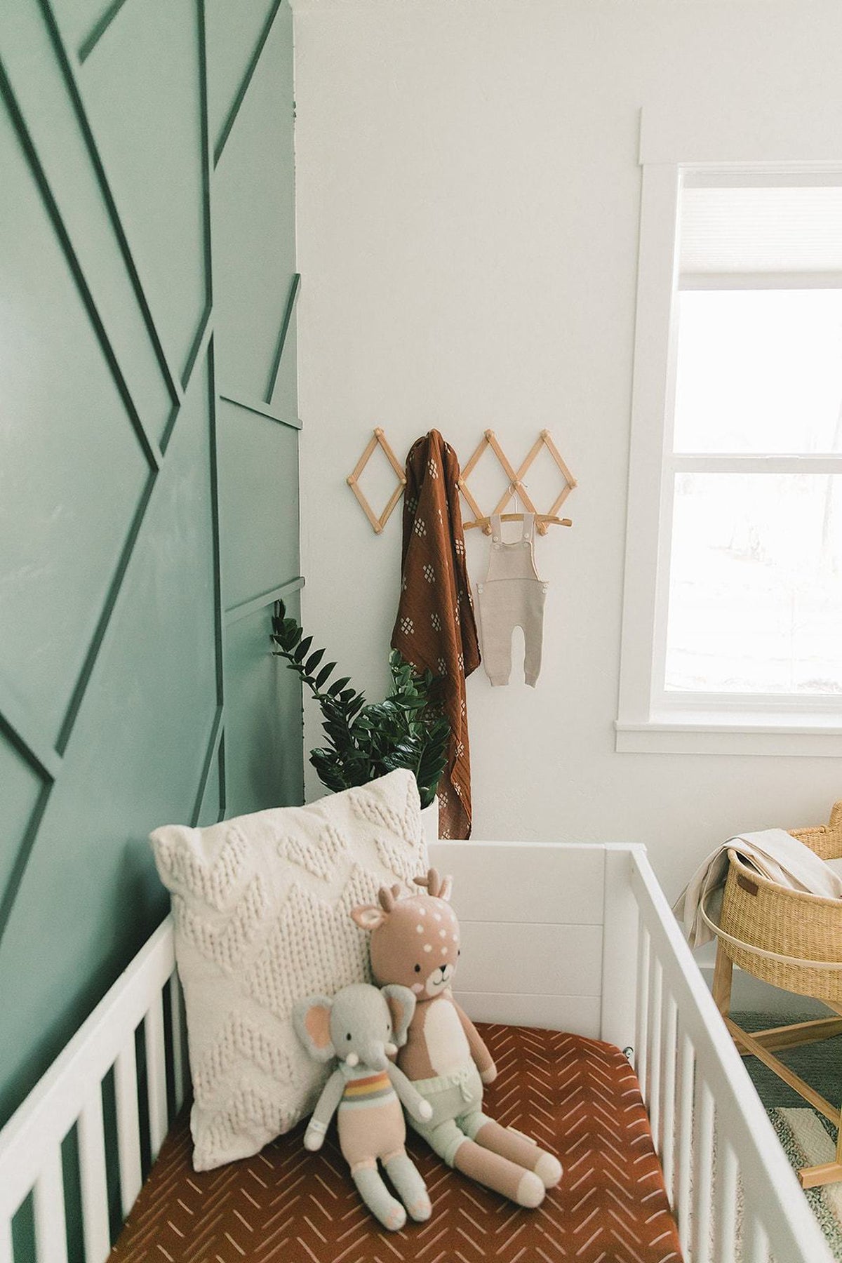 A nursery with a white crib, rust muslin sheet, plush toys, and geometric green wall design.