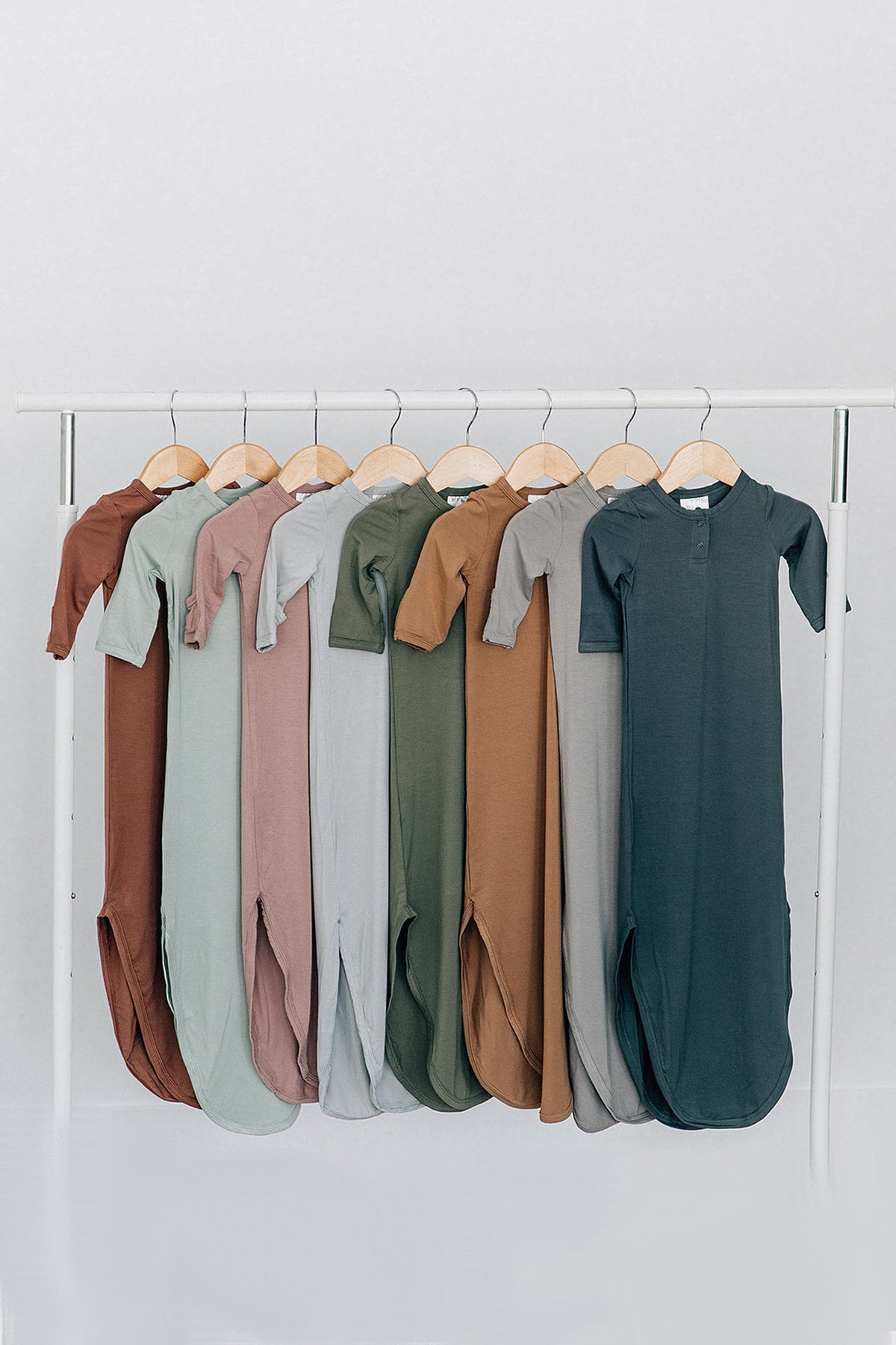A row of baby knot gowns in various earth tones hangs on wooden hangers against a light background.