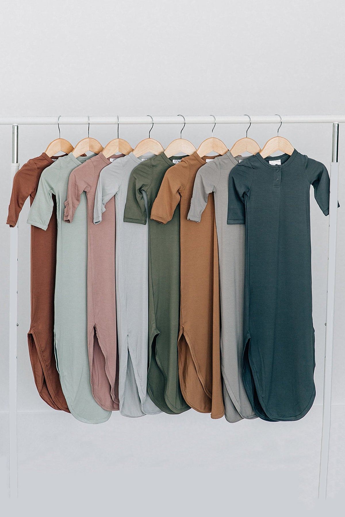 Assorted baby gowns in earthy tones hang on wooden hangers, showcasing soft bamboo fabric and shoulder flaps.