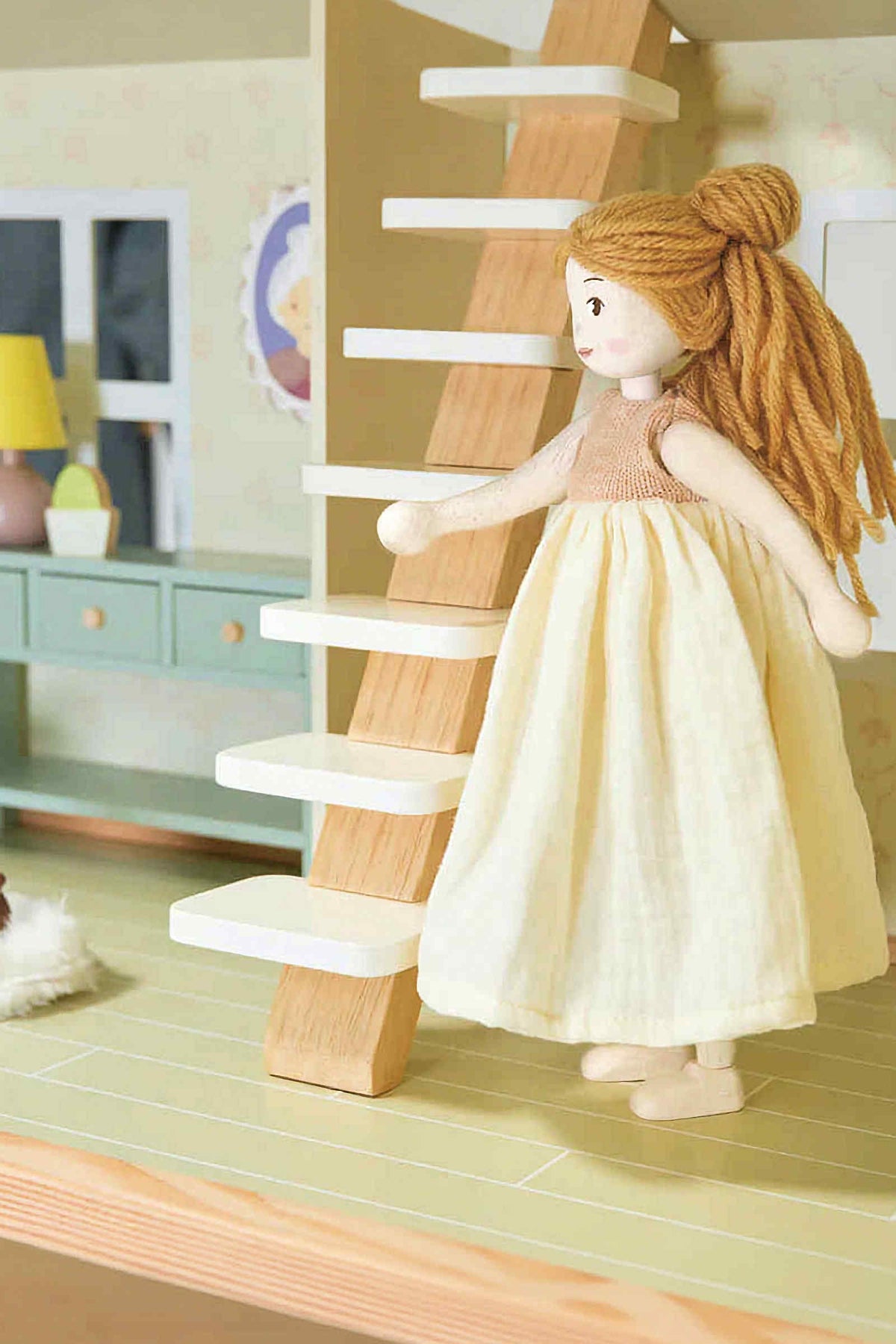 A handcrafted wooden doll with blonde yarn hair and a cream dress ascends a light wooden staircase in a dollhouse setting. The doll is designed for imaginative play and sustainability.