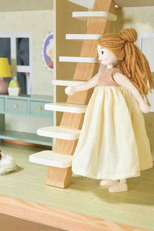 A handcrafted wooden doll with blonde yarn hair and a cream dress ascends a light wooden staircase in a dollhouse setting. The doll is designed for imaginative play and sustainability.