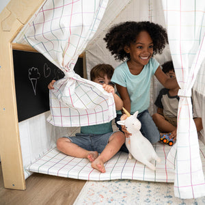 Two children play inside a cozy wooden playhouse with a checkered fabric design. One child holds a plush unicorn while they enjoy their imaginative space together.