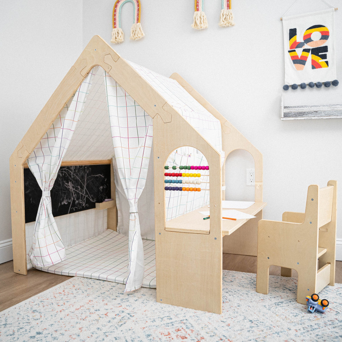 A wooden playhouse with a white grid-patterned canopy features a chalkboard and a colorful abacus. Adjacent is a matching desk and chair, creating a cozy, creative space for children.