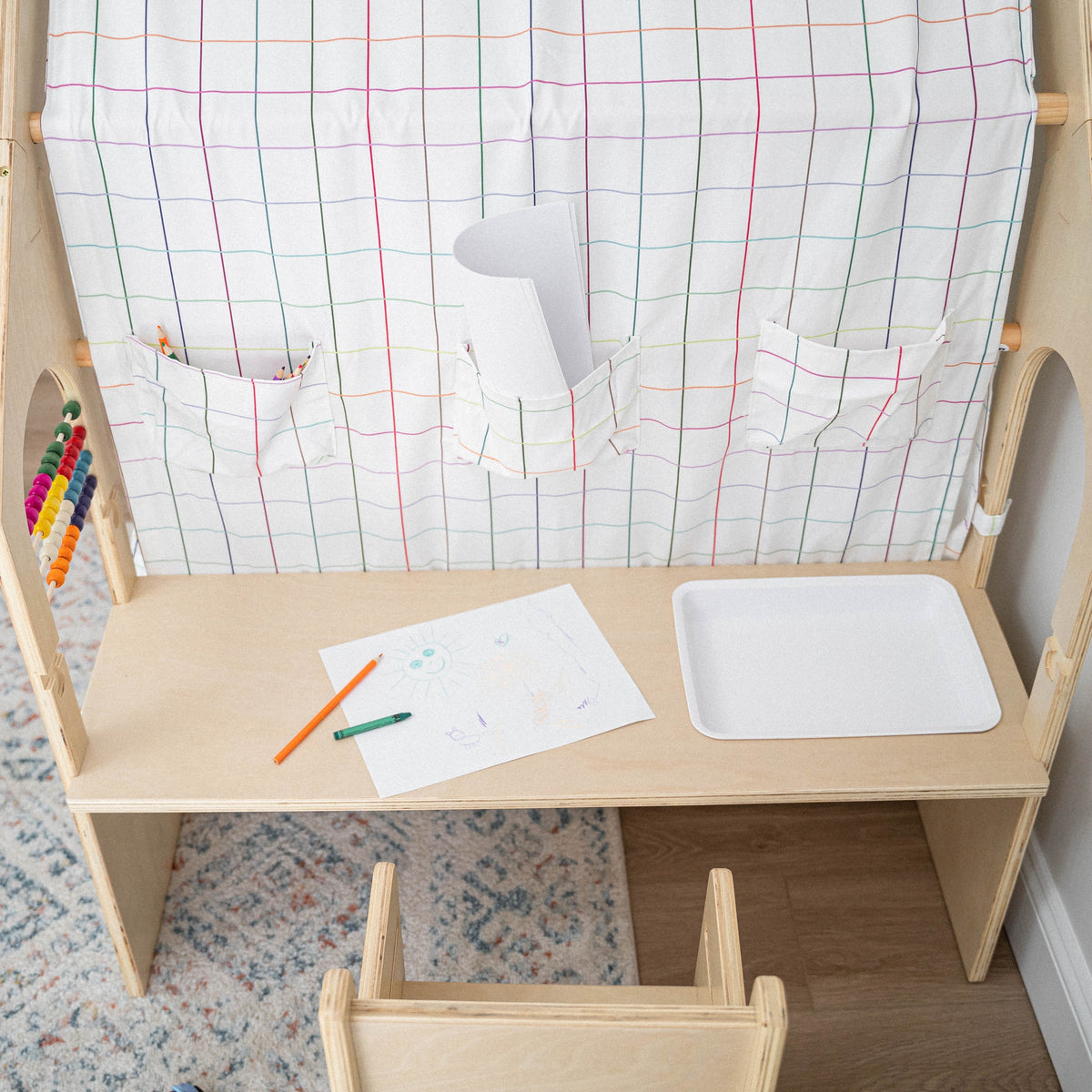 A wooden desk features a colorful grid-patterned fabric backdrop with pockets for art supplies. The scene includes a drawing pad and colored pencils, promoting creativity and play for children.
