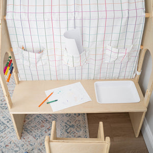 A wooden desk features a colorful grid-patterned fabric backdrop with pockets for art supplies. The scene includes a drawing pad and colored pencils, promoting creativity and play for children.