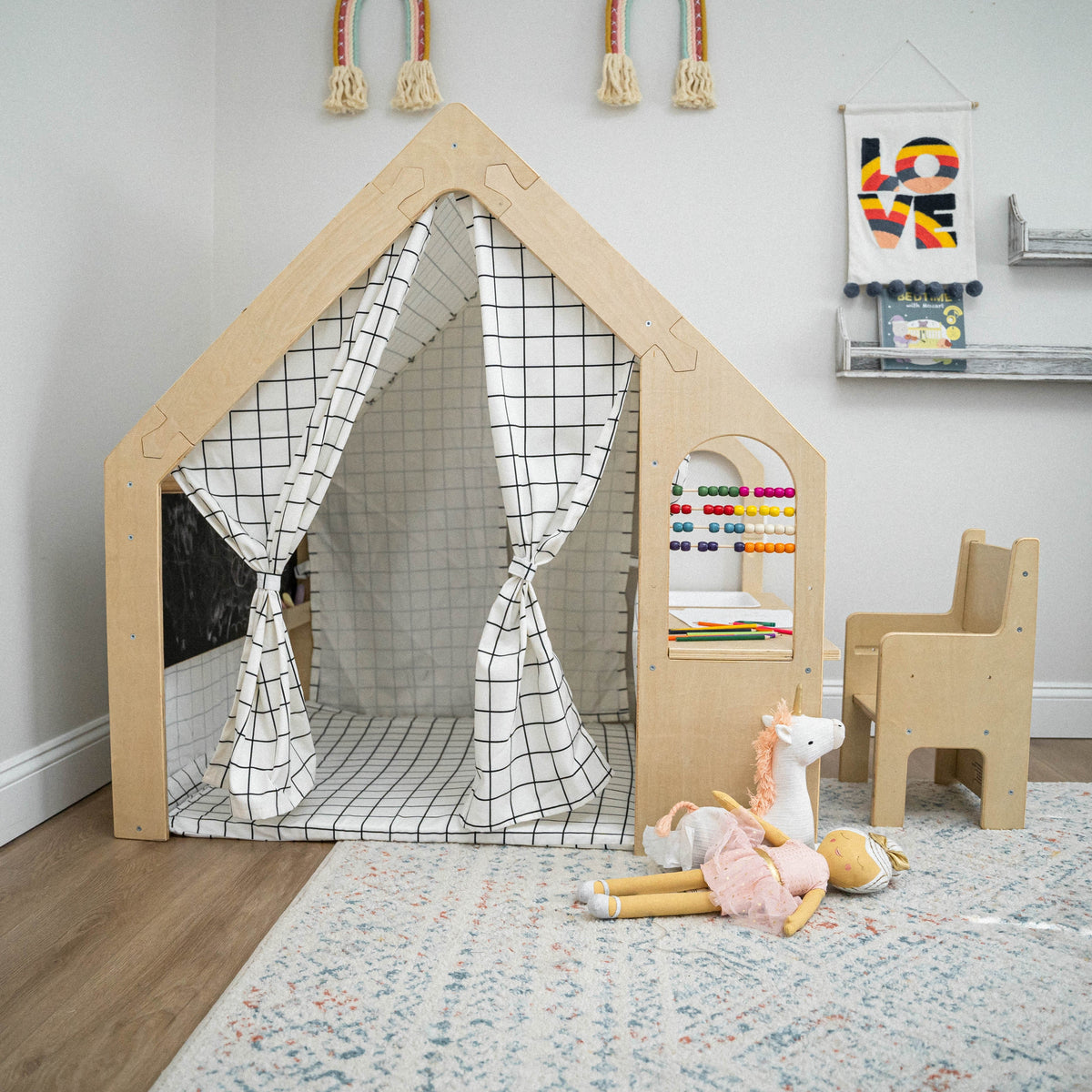 A wooden playhouse with a grid-patterned curtain features an adjacent sensory desk with colorful beads and a chair. Plush toys are scattered on a soft area rug nearby.