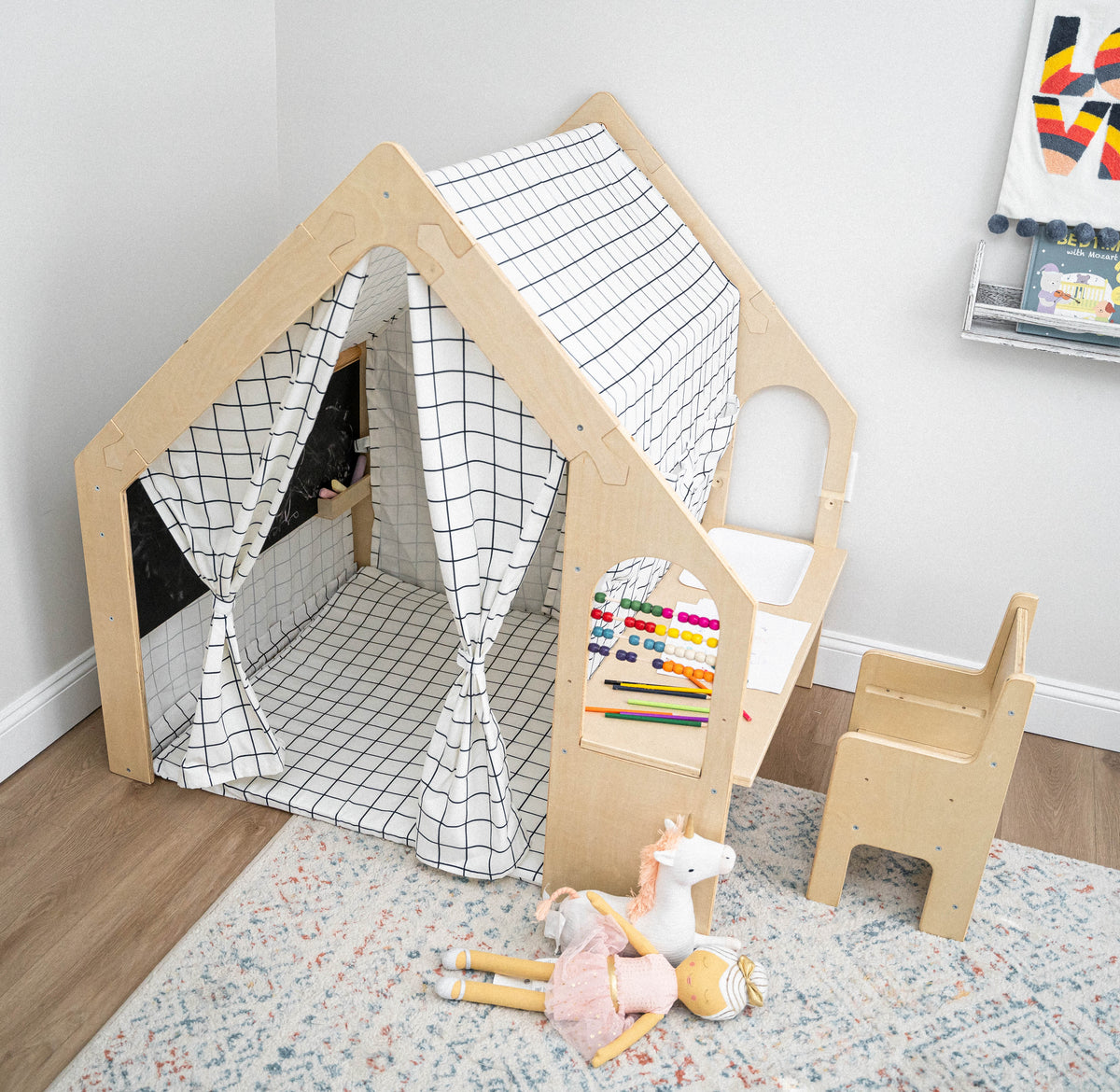 A wooden playhouse features a grid-patterned canopy and a small desk with a chair. Colorful beads and a plush toy are placed nearby, creating a cozy play environment for children.