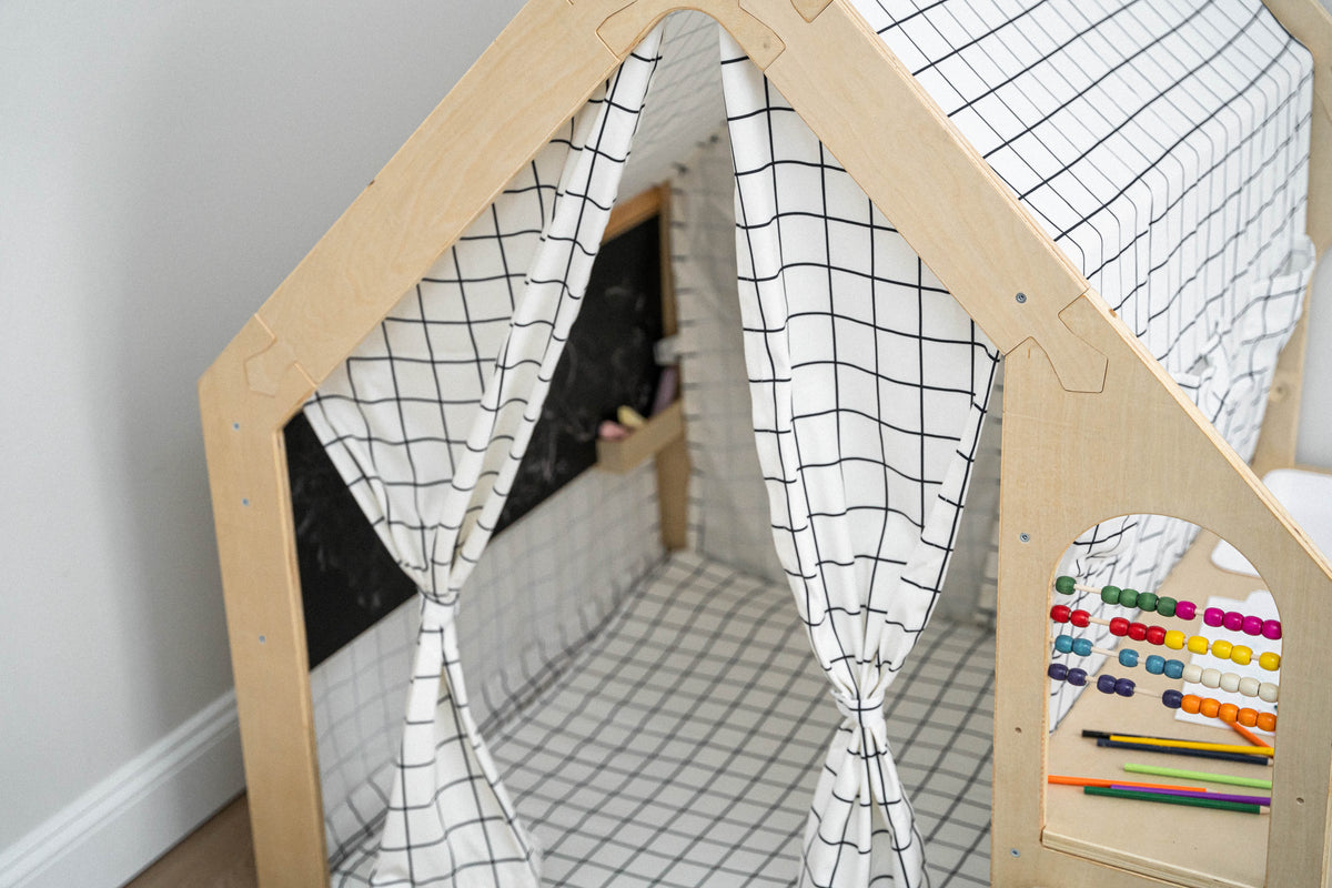 A cozy wooden playhouse features a black and white grid design, with curtains and a chalkboard inside. Colorful abacus beads and crayons are visible nearby, promoting creativity and learning.