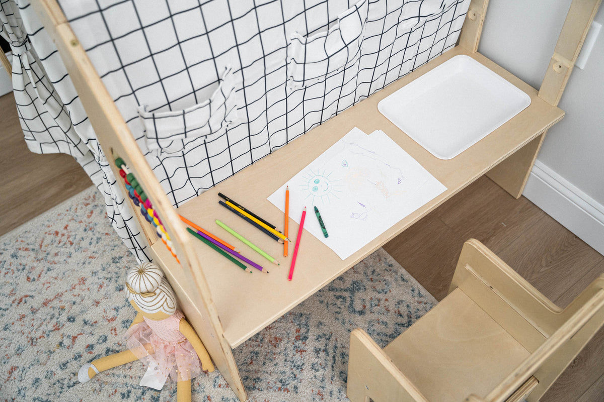 A wooden desk with a simple design features a white drawing surface, surrounded by colorful art supplies. Nearby, a small chair and a doll add to the inviting play space.