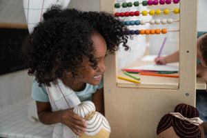 A smiling child with curly hair holds a plush toy while playing in a wooden playhouse. Brightly colored beads on an abacus and art supplies are visible in the background.