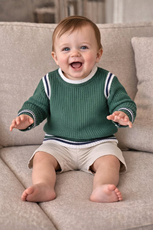 A smiling baby boy sits on a beige couch, wearing a green organic cotton crew neck sweater with sporty white and navy stripes.