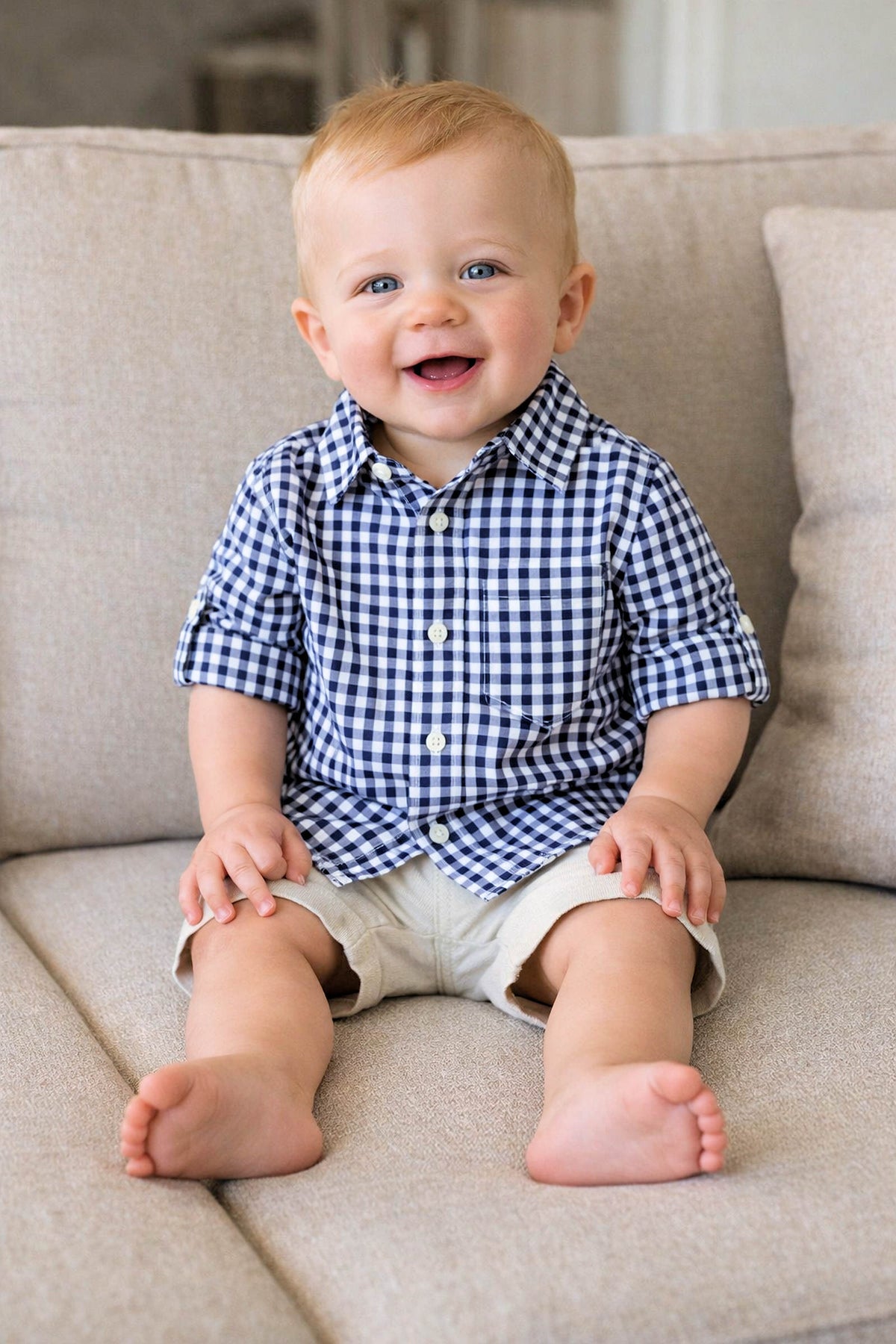 A cheerful baby sits on a light beige sofa, wearing a navy gingham button-down shirt and light shorts. The soft cotton fabric and classic style add a touch of charm.