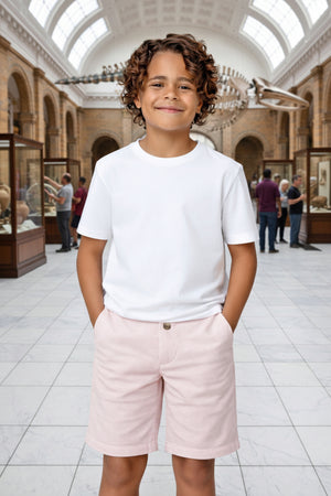A smiling boy wears a white t-shirt and pink seersucker shorts, standing in a museum with exhibits in the background.