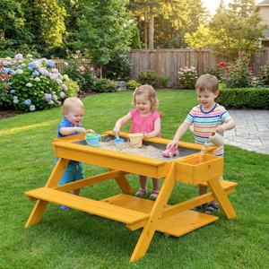 Three children play joyfully at a bright yellow outdoor picnic and sand table, scooping sand with colorful buckets in a lush garden setting surrounded by blooming flowers and greenery.