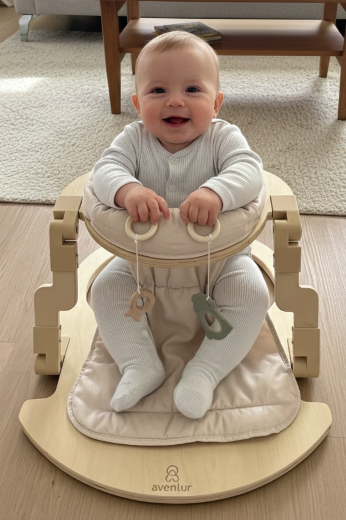 A happy baby sits in a light wooden activity seat with a soft, padded cushion. The seat features natural materials and is designed for safe and enjoyable playtime.