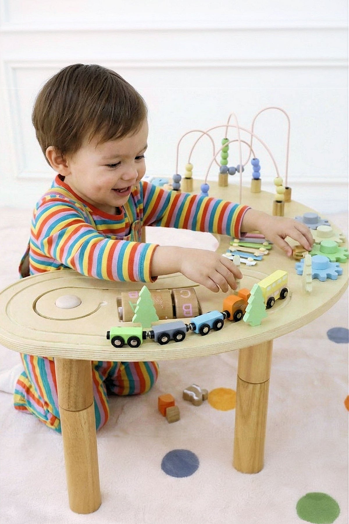A smiling child in colorful striped pajamas plays with a wooden multi-activity sensory table featuring trains and various interactive elements.