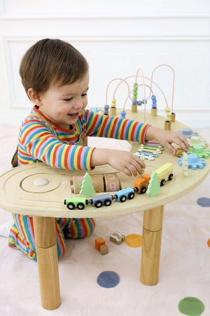 A smiling child in colorful striped pajamas plays with a wooden multi-activity sensory table featuring trains and various interactive elements.