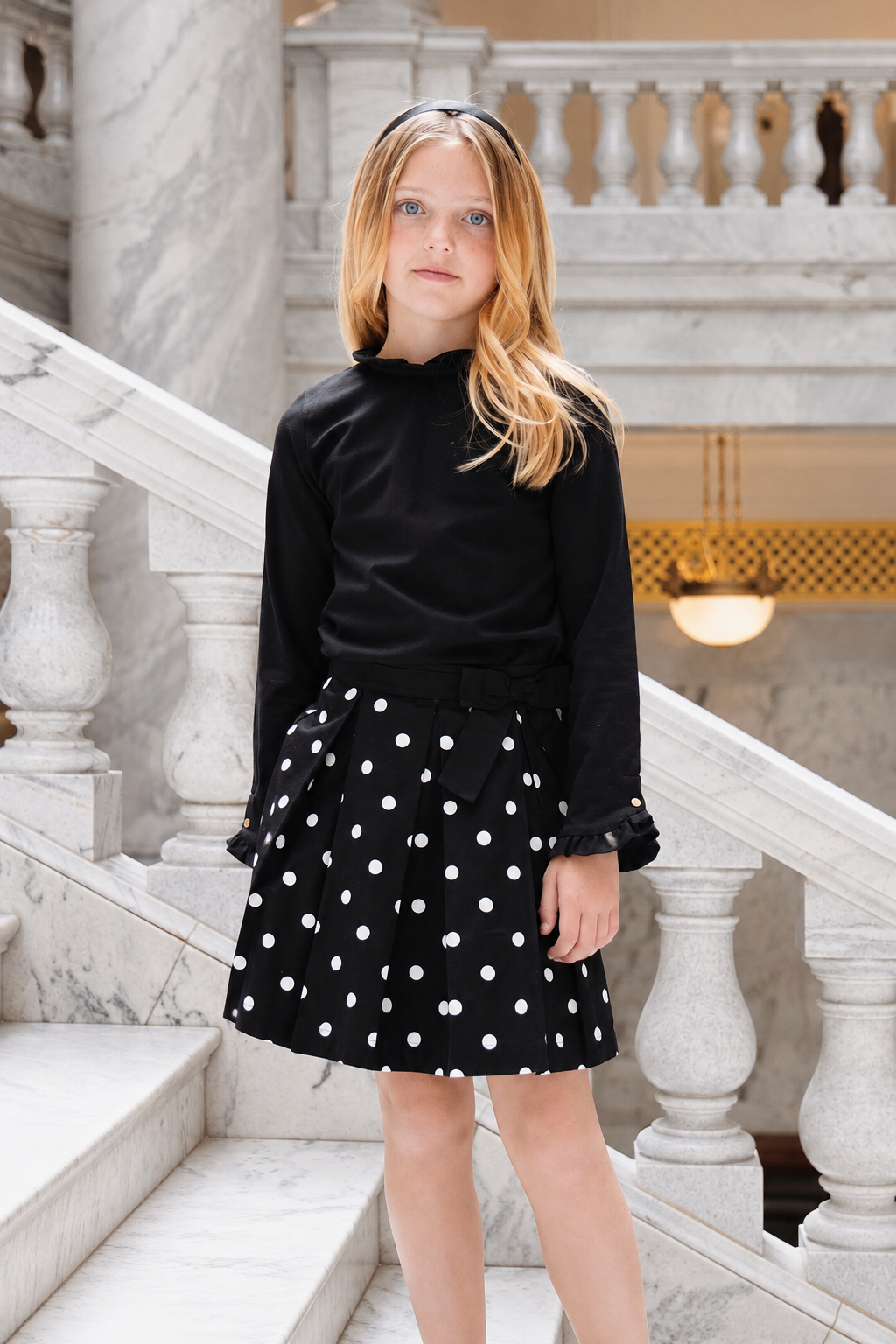 A young girl stands on a marble staircase, wearing a black long-sleeve top and a black pleated skirt adorned with white polka dots.