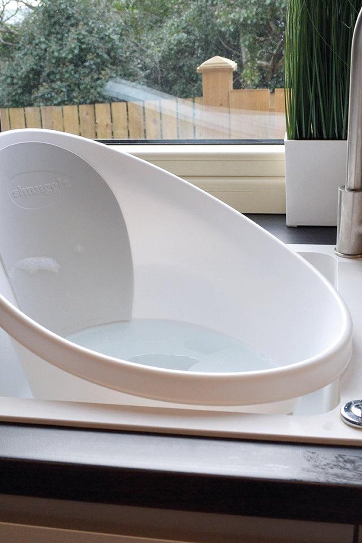 White Shnuggle baby bath with ergonomic design sits on a kitchen counter near a window and potted plant.