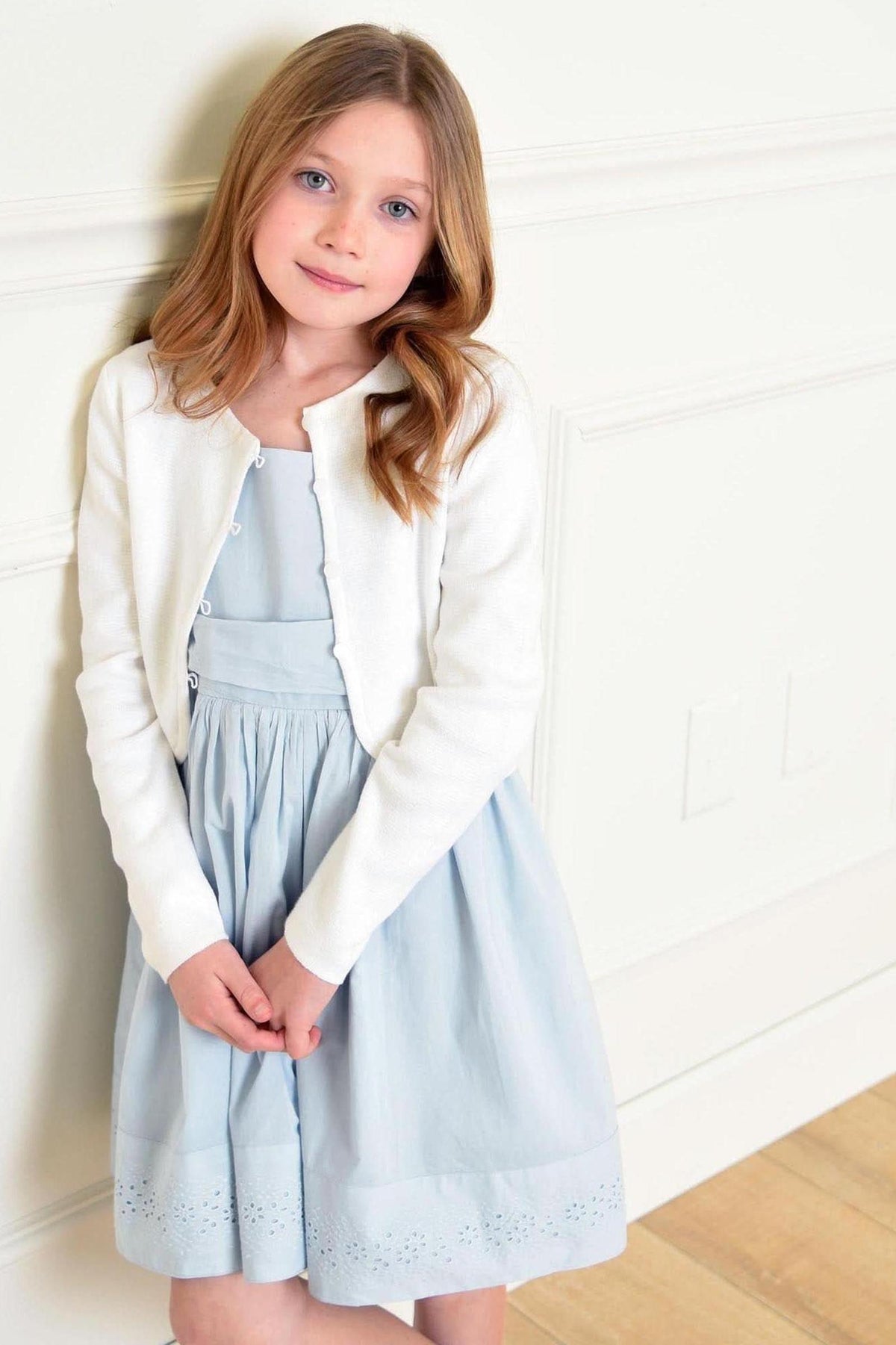 A young girl in a white cropped cardigan poses in front of a light-colored wall, wearing a light blue dress.