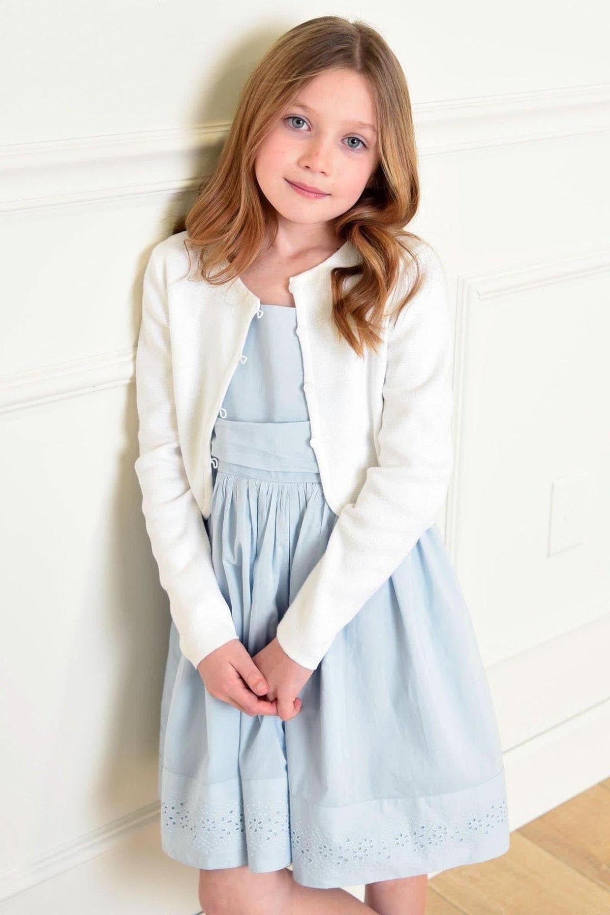 A young girl wearing a white cardigan and a light blue dress stands against a neutral wall, smiling softly.