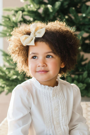 A young girl with curly hair wears a white ruffled blouse, smiling softly in front of a green backdrop.
