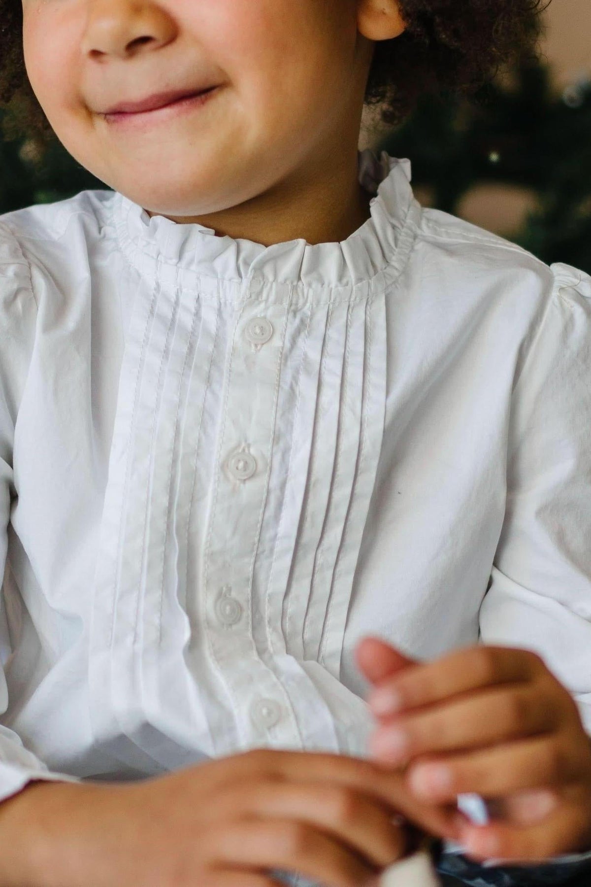 A young child smiles while wearing a white blouse with a ruffled neckline and pintucks, showcasing delicate button details.