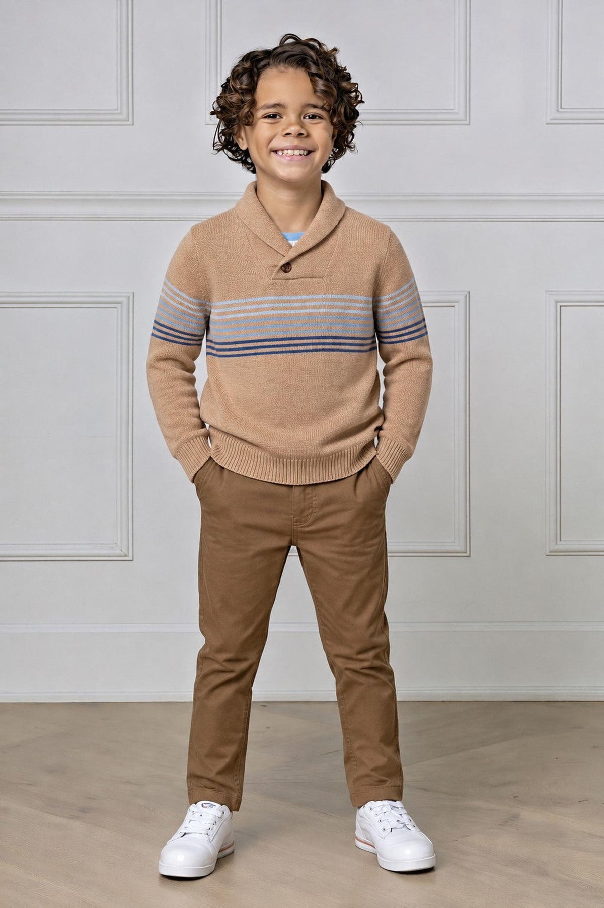 A young boy smiles while wearing a camel-colored organic cotton shawl collar sweater with blue stripes, paired with brown pants and white sneakers, standing in a bright, neutral-toned setting.