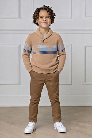 A young boy smiles while wearing a camel-colored organic cotton shawl collar sweater with blue stripes, paired with brown pants and white sneakers, standing in a bright, neutral-toned setting.