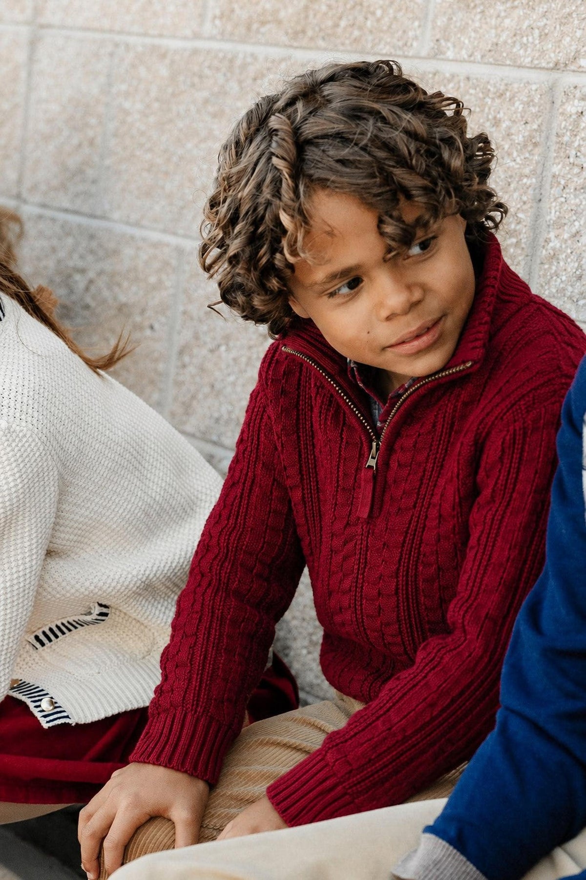 A young boy with curly hair wears a rich oxblood cable-knit sweater, featuring a half-zip design. He sits casually beside others against a textured wall, showcasing a cozy, stylish look.