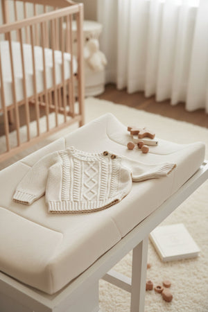 A cozy ivory cable knit sweater is displayed on a soft changing pad, surrounded by wooden toys.