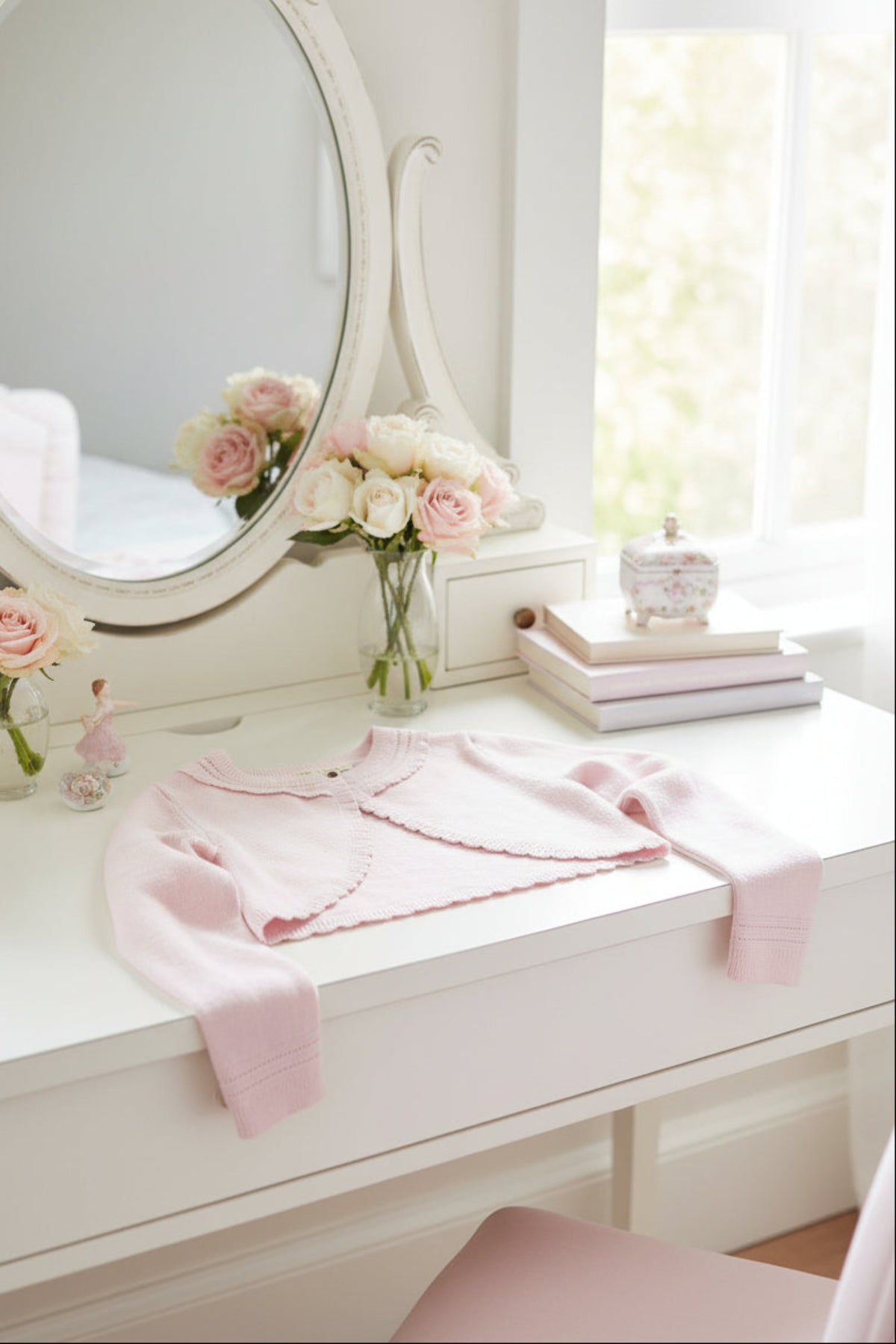 A soft pink cropped cardigan rests on a white desk, surrounded by delicate flowers and a decorative mirror.