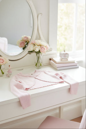 A soft pink cropped cardigan rests on a white desk, surrounded by delicate flowers and a decorative mirror.