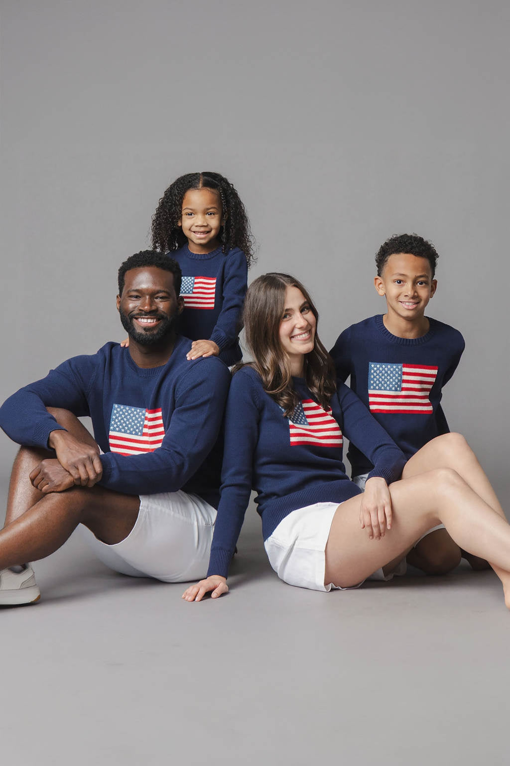 A family of four poses together, wearing matching navy sweaters featuring an American flag design. The soft, organic cotton sweaters are ideal for coordinated family moments and celebrations.