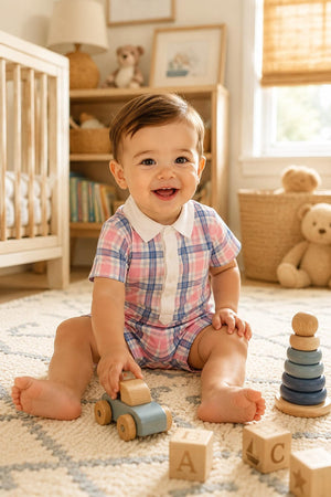 A smiling baby sits on a patterned rug in a cozy nursery, wearing a fresh pink and blue plaid romper. Wooden toys, including a car and stacking rings, are nearby.