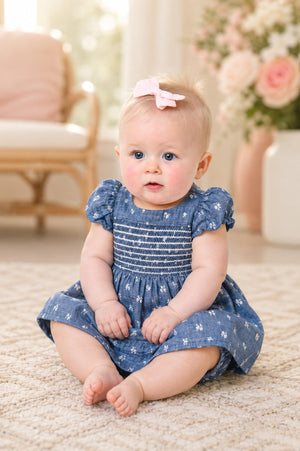 A baby sits on a soft rug, wearing a blue chambray dress with a smocked bodice and delicate white embroidery. The background features soft floral decor.