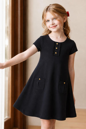 A young girl smiles while wearing a black organic cotton A-line dress featuring a ruffle placket and two front pockets, showcasing a classic and timeless style suitable for various occasions.
