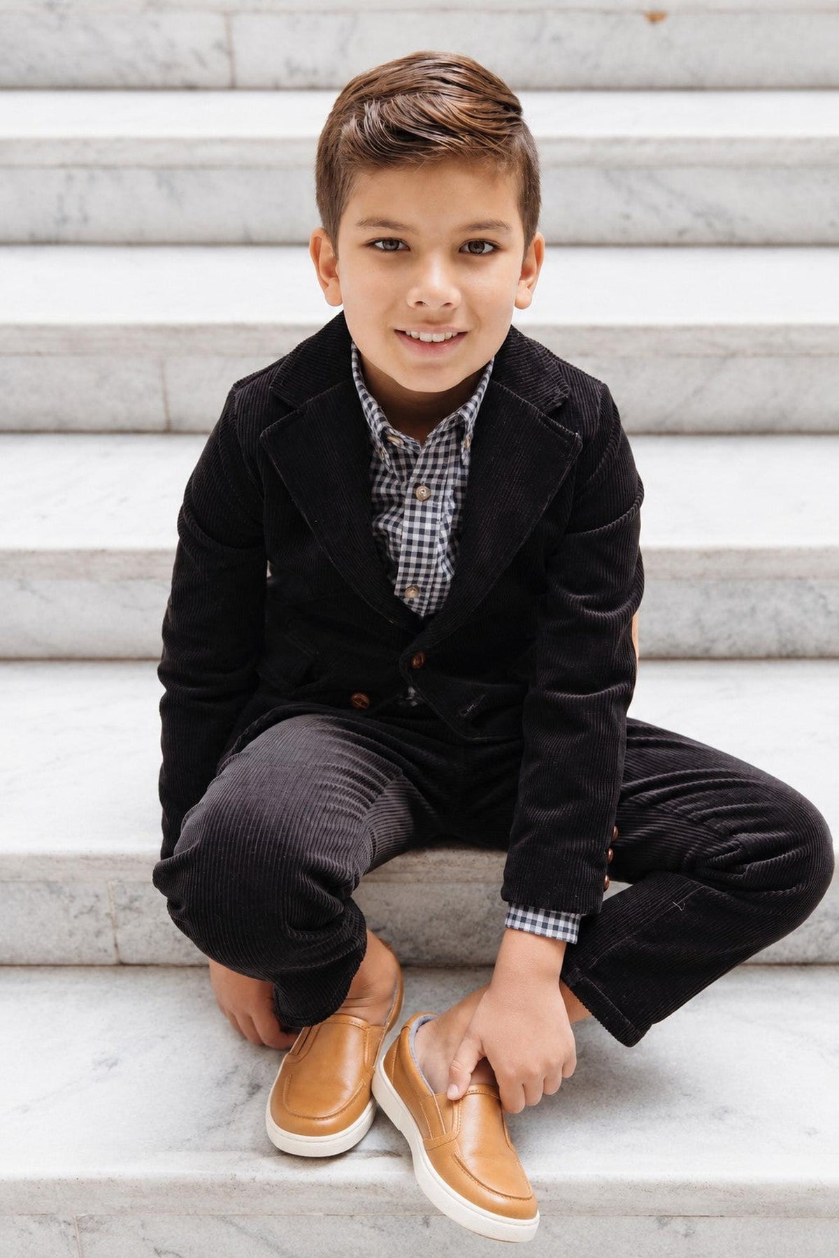 A young boy sits on marble stairs, wearing a black corduroy suit with a black gingham button-down shirt and brown slip-on shoes, showcasing a stylish.