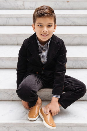A young boy sits on marble stairs, wearing a black corduroy suit with a black gingham button-down shirt and brown slip-on shoes, showcasing a stylish.